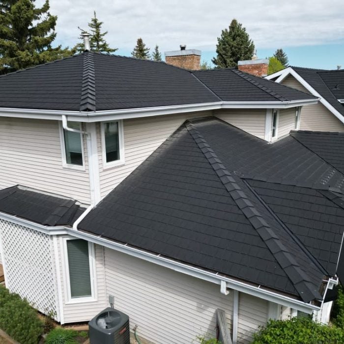 Detailed aerial drone photograph looking down at a complex, multi-level residential home with light beige siding. The prominent roof is a multi-pitched structure covered in high-quality, dark grey synthetic rubber roofing that replicates the appearance of premium slate tiles. The house features multiple windows with white trim, white gutters, a large white lattice section, and an A/C unit on a pad, all set within a suburban landscape of mature pine and deciduous trees under a cloudy sky. This image illustrates a prime example of expert residential rubber roofing installation or repair.